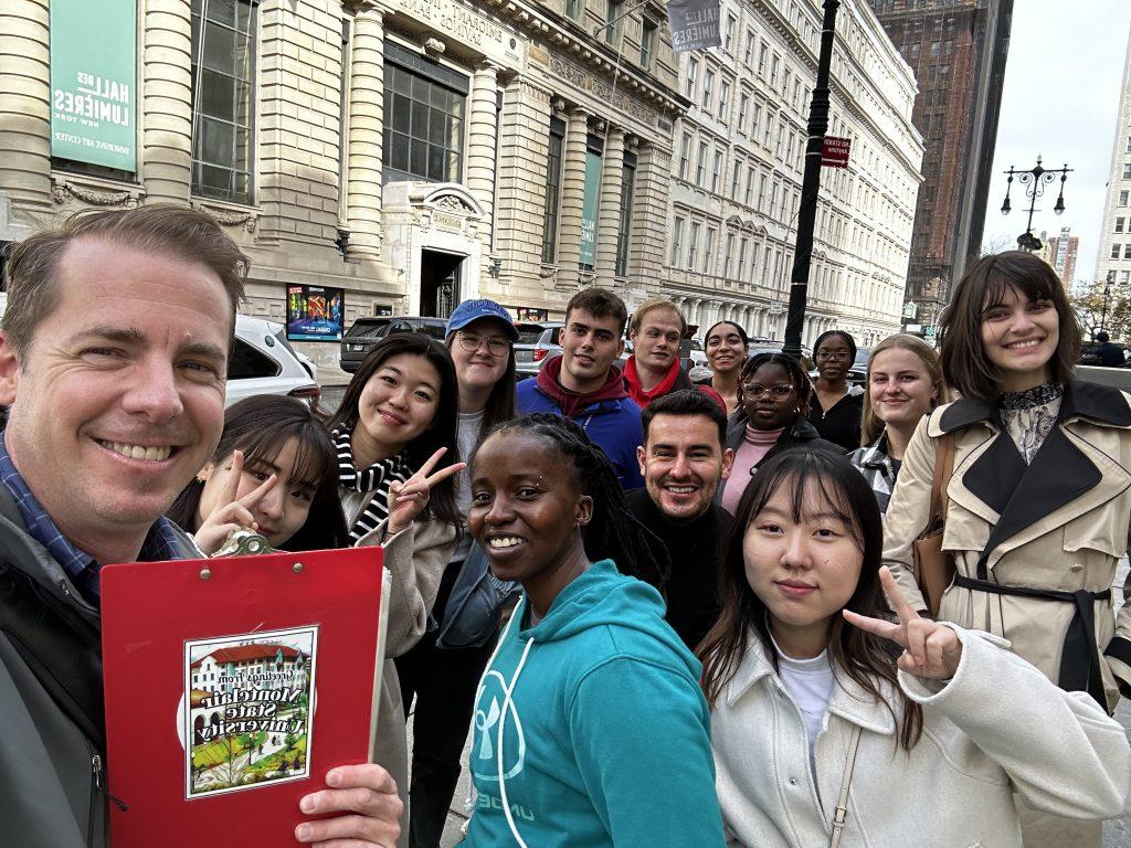 Group of students from different countries and cultures stand together in New York City with a man holding a red clipboard that says "Montclair State University" on the back